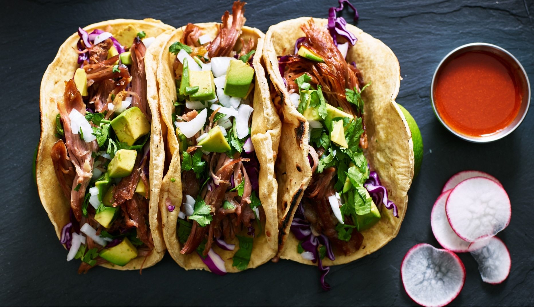 A top-down view of three soft corn tortilla tacos filled with shredded, slow-cooked meat and topped with diced avocado, chopped white onion, fresh cilantro, and shredded purple cabbage. The tacos are arranged side by side on a dark slate serving surface. To the right is a small bowl of red salsa, with lime wedges and sliced radishes nearby. The colorful toppings contrast with the dark background, creating a vibrant, appetizing presentation.