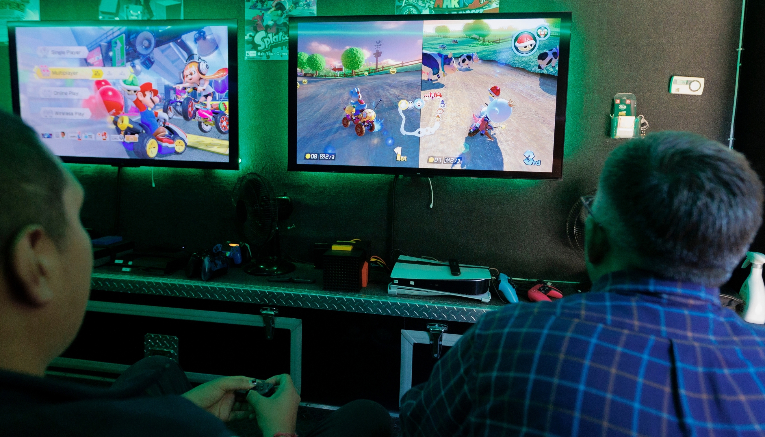 Two people sit facing two large flat-screen TVs while playing a split-screen racing video game in a dimly lit gaming room with green accent lighting. One person in the foreground holds a game controller, while the other is seen from behind wearing a blue plaid shirt. The left screen displays the game's multiplayer menu, and the right screen shows an active race with two players driving on a countryside track. Below the screens are gaming consoles, controllers, and small fans on a counter, with posters decorating the wall in the background.