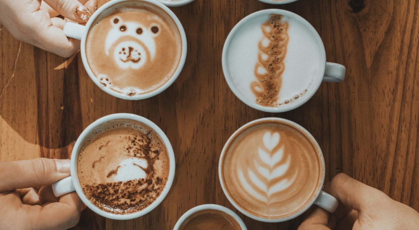 Overhead view of a wooden table with four cups of coffee featuring different latte art. The top-left cup has a bear face design and is being held by a person with floral nail art. The top-right cup shows a vertical chain-like foam pattern. The bottom-left cup has a landscape-style design resembling a tree beside a hill, with cocoa powder sprinkled around the edge, and is held by another person. The bottom-right cup features a classic leaf (rosetta) latte art pattern and is being held by a third person. A small cup is partially visible at the bottom center, and the warm lighting highlights the cozy café setting.