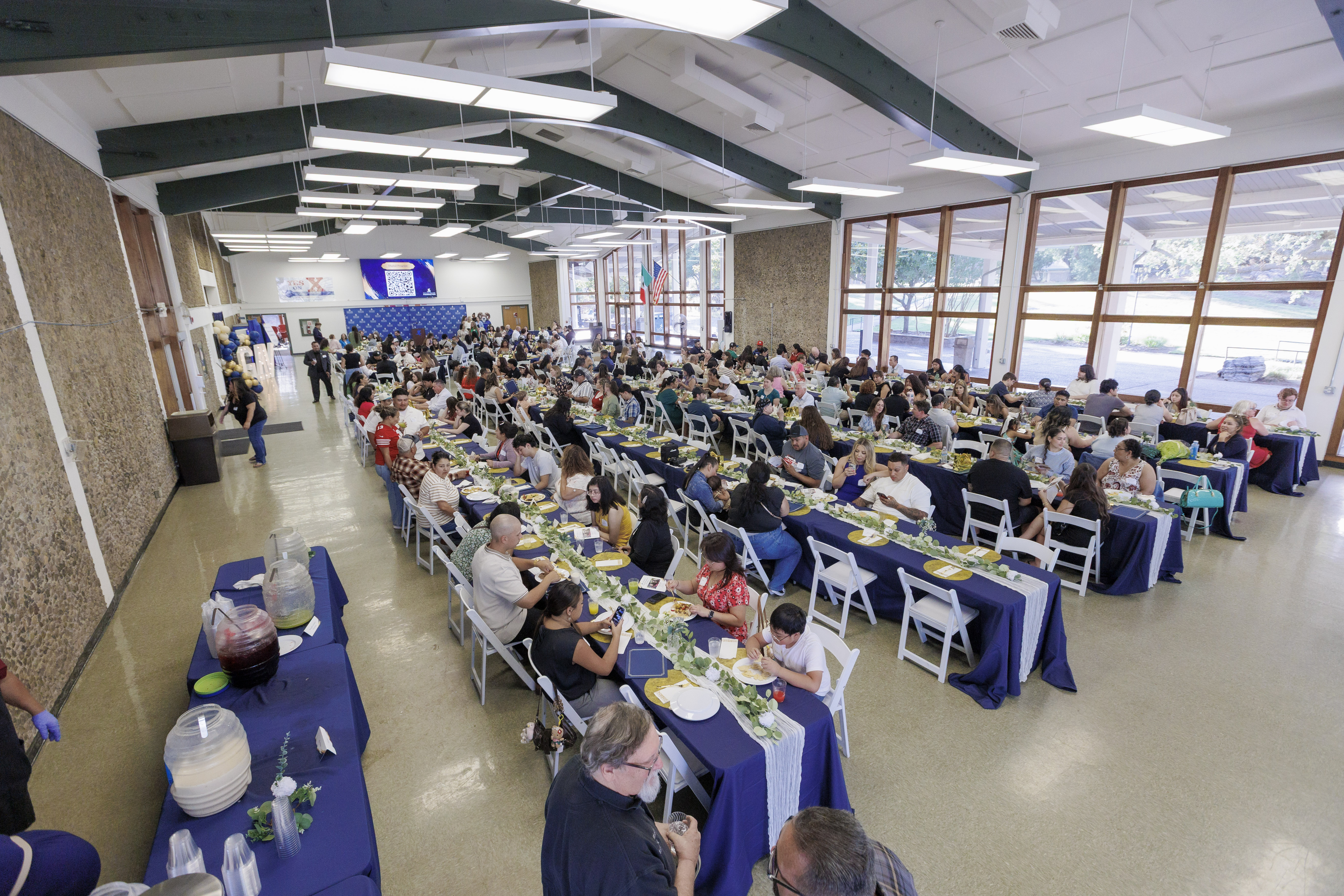A large group of people gathers for a scholarship awards dinner in a spacious hall, seated at long tables decorated with blue tablecloths, greenery, and place settings.