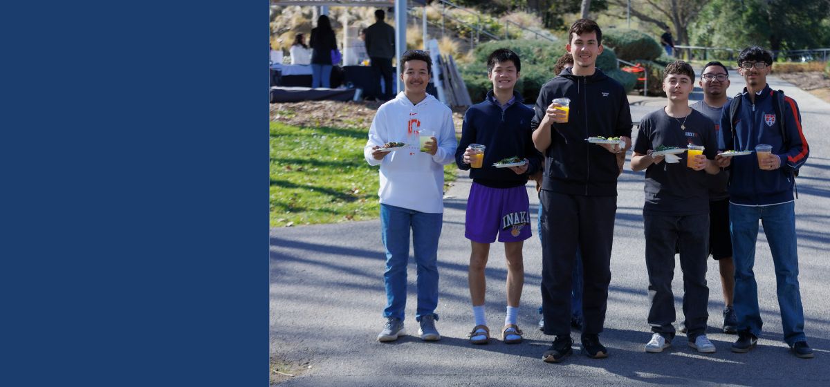 Six students stand outdoors on campus holding plates of food and drinks, smiling toward the camera.