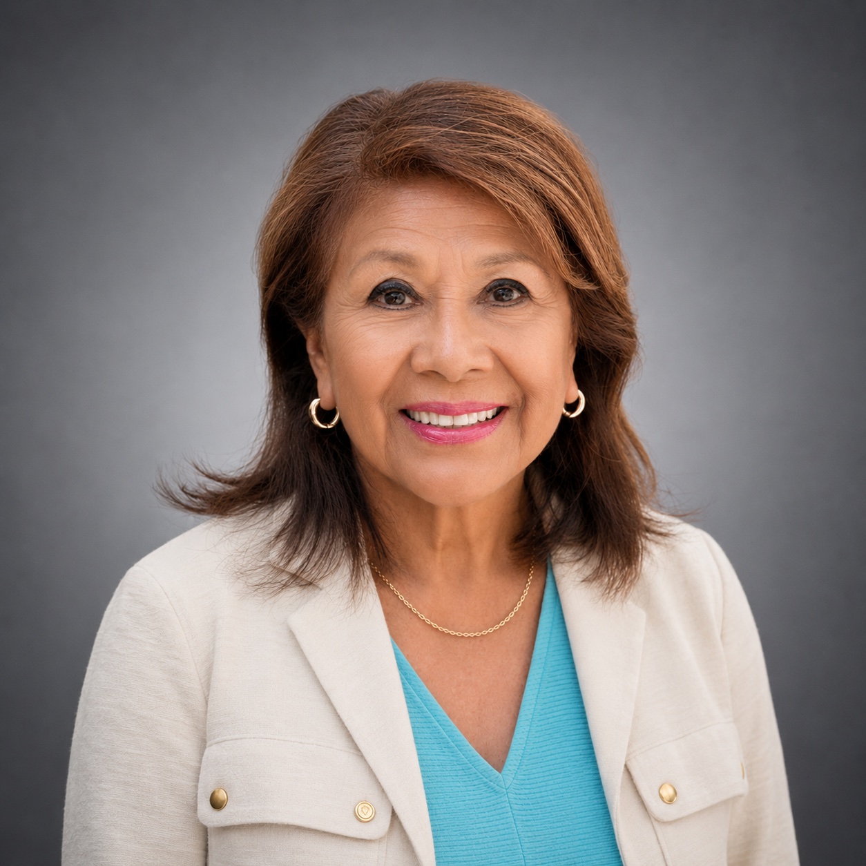 Professional headshot of a smiling Evelia Morales Rosso looking directly at the camera against a gray studio background. They are wearing a light cream blazer over a turquoise blouse, along with small gold hoop earrings and a delicate gold necklace. The portrait is cropped from the chest up and features soft, professional lighting.