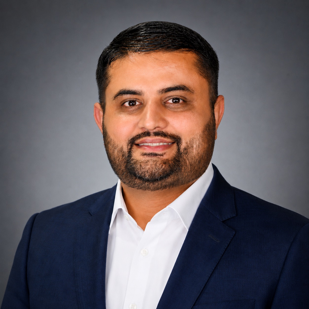 Professional headshot of Manny Bhandal smiling at the camera against a gray background. He is wearing a navy blue suit jacket over a white collared shirt. The portrait is cropped from the chest up and features studio lighting, creating a polished and professional appearance.