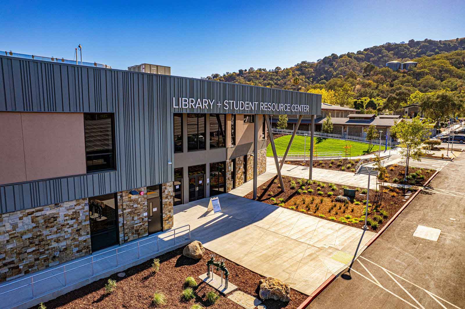 Exterior view of Gavilan College’s Library and Student Resource Center, featuring a modern two-story building with blue-gray metal siding, stone accents, and large windows. A wide concrete entrance plaza, accessible ramp, landscaped garden beds, and parking spaces surround the building. Green hills and a clear blue sky are visible in the background.