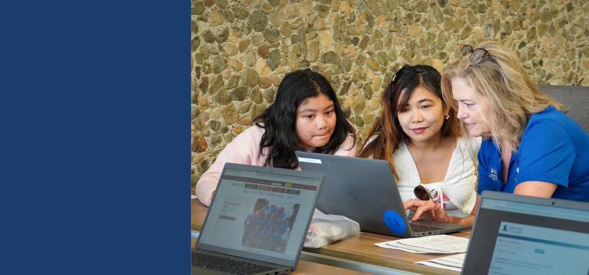 Two students work with a Gavilan College staff member at a table, looking at a laptop while other laptops and materials are visible in the foreground.