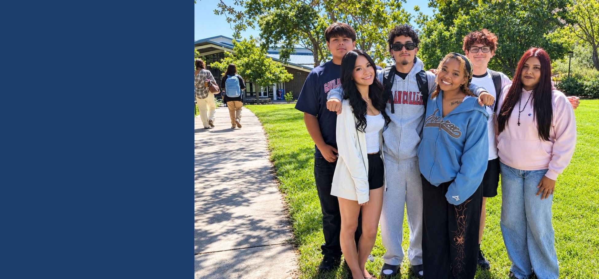 Six college students stand together on a sunny campus lawn, smiling at the camera. They are gathered closely with their arms around one another, with trees, grass, a campus building, and a walkway in the background.
