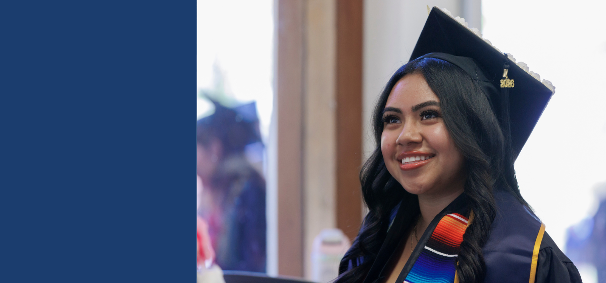 A smiling graduate wearing a black cap and gown and a colorful graduation stole looks toward the camera. She has long dark hair and is pictured indoors, with other graduates blurred in the background.