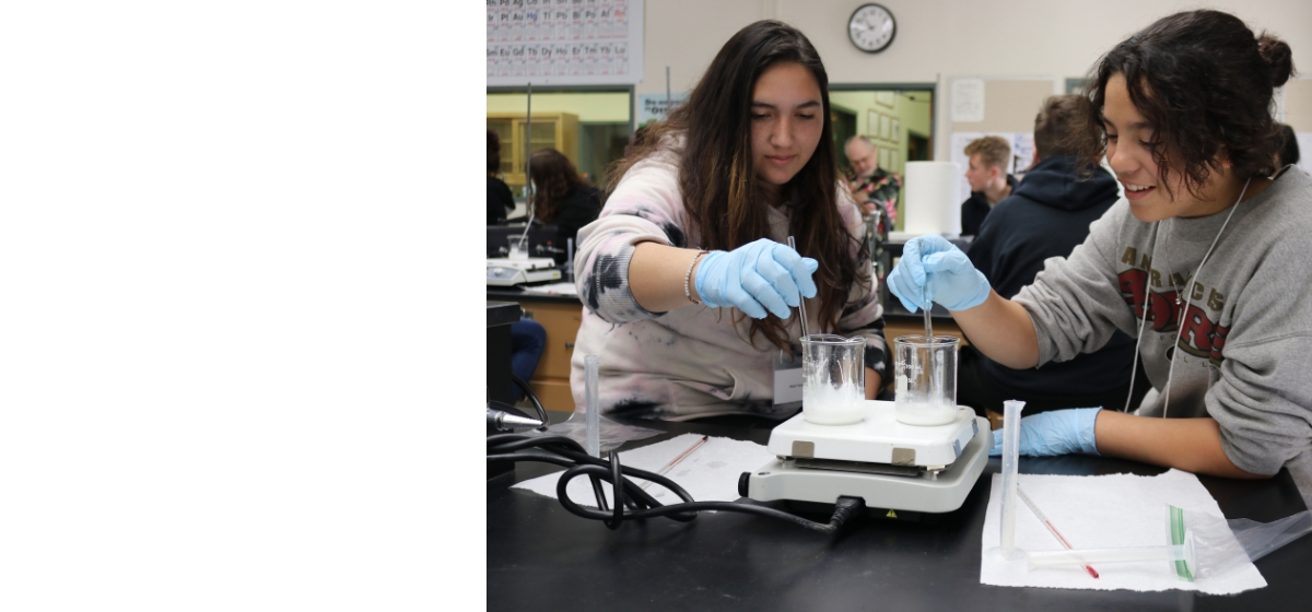 Two female students wearing gray sweatshirts stirring glass beakers on a scale in a classroom 