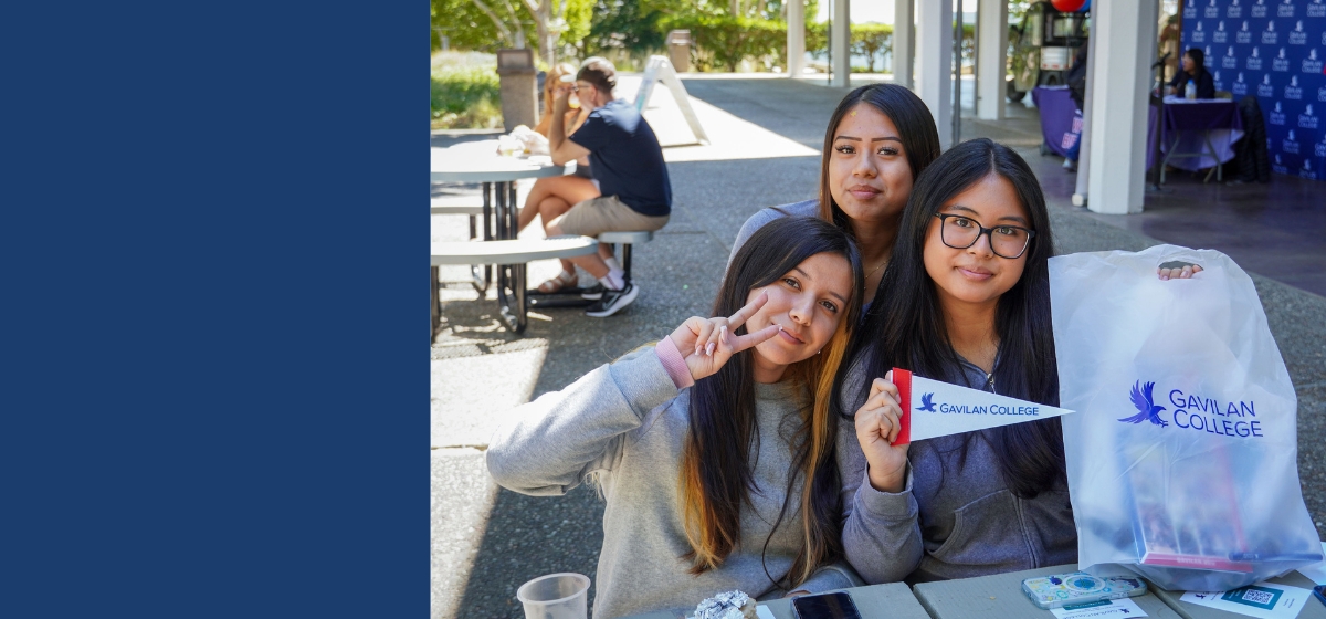 Three female students sitting outside of the Gilroy Student Center holding up a little Gavilan College flag and a Gavilan College plastic bag as one flashes a piece sign with her fingers