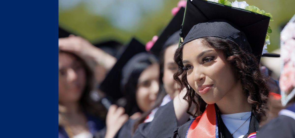 Image of African American female graduate with curls and a red sash look off to the side. Blurry grads can be seen in the background.