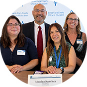 Four people pose together for a photo, with one woman seated in front of a backdrop featuring Santa Clara County branding. The group is smiling and wearing name badges.